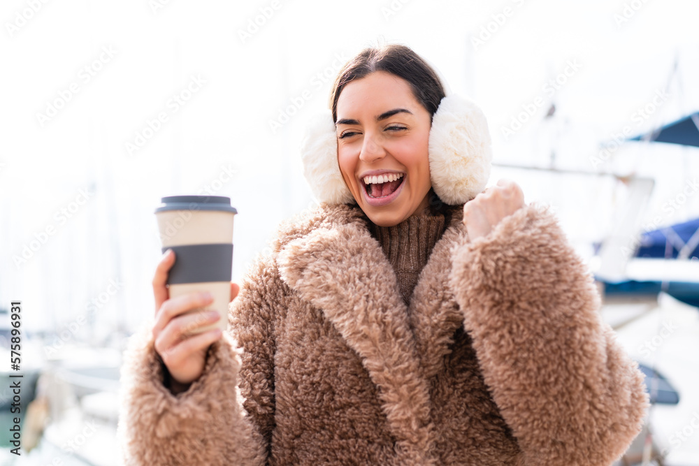 Young woman wearing winter muffs and holding take away coffee at outdoors celebrating a victory Young woman wearing winter muffs and holding take away coffee at outdoors celebrating a victory