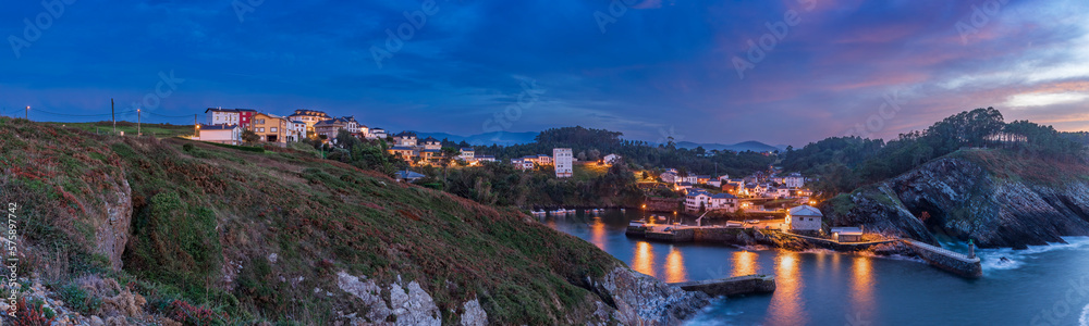 Fototapeta premium View of the small town of Viavelez with reflections of lanterns in the harbor on the coast of Spain and where the moon breaks through the sunset clouds in the evening sky, Asturias, Spain.