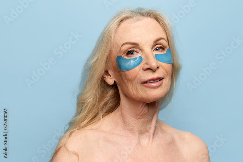 Foto Beautiful middle-aged woman with well-kept healthy skin posing with under eye patches over blue studio background