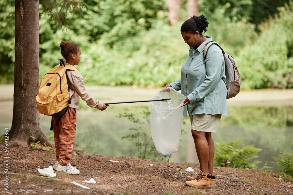 Side view portrait of black mother and daughter helping cleaning nature together and picking up trash by river, copy space  - obrazy, fototapety, plakaty 