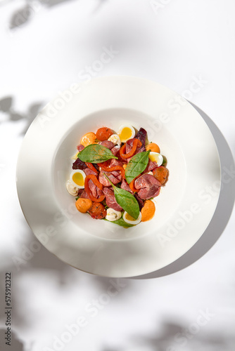 Salad with roast beef, vegetables and eggs on white table with harsh shadows. Steak salad in modern style with shadows of leaves. Flashy food. Meat salad with pastrami in summer menu.