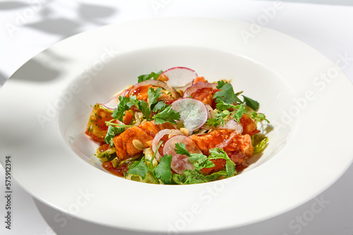 Elegant salad with baked salmon in sauce with vegetables on white table with harsh shadows. Salmon salad with radish on white background with shadows of leaves. Summer salad with roasted trout.