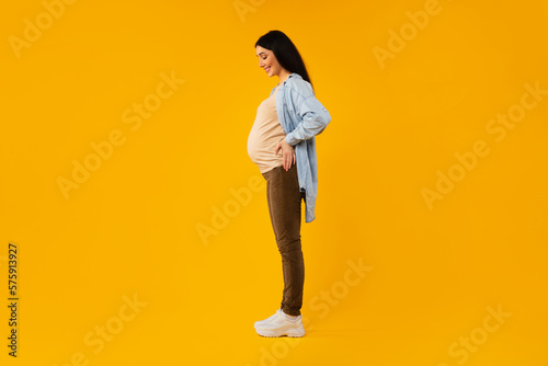 Profile shot of pregnant woman posing on yellow background, looking at belly and smiling, enjoying awaiting time