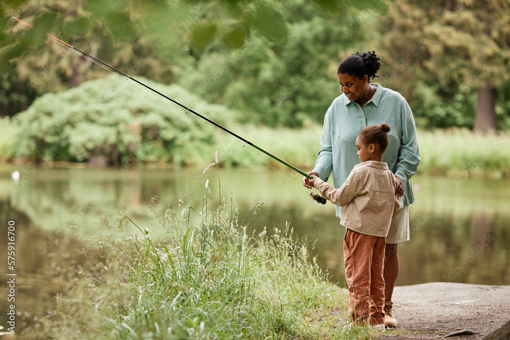 Side view portrait of caring black mother teaching little girl fishing by beautiful forest lake, copy space  - obrazy, fototapety, plakaty 