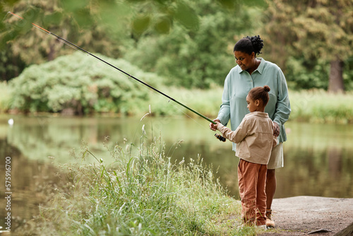 Side view portrait of caring black mother teaching little girl fishing by beautiful forest lake, copy space 