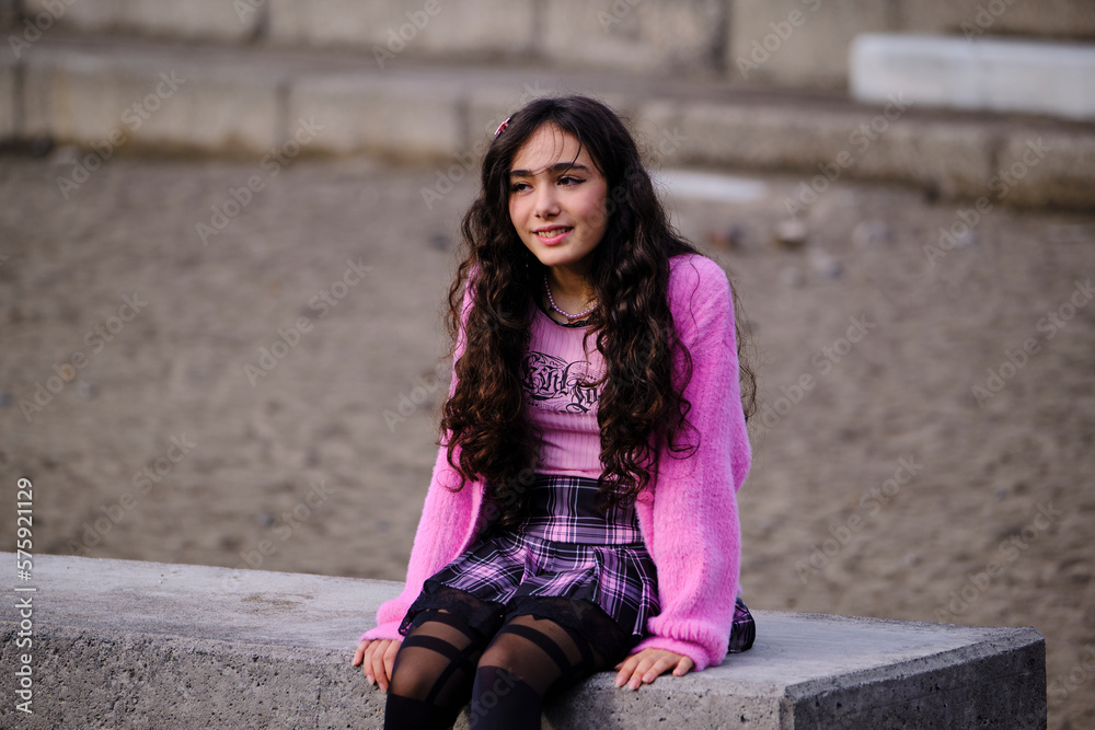 Fototapeta premium Profile portrait of teenage girl posing dressed in gothic style in black and pink colours sitting on a wall