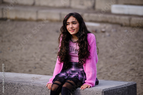 Profile portrait of teenage girl posing dressed in gothic style in black and pink colours sitting on a wall