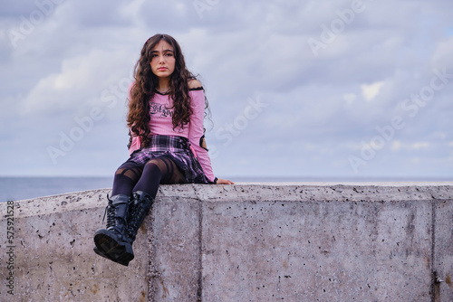 Portrait of smiling teenage girl with gothic and boyish look posing sitting on a wall in front of the sea. 
