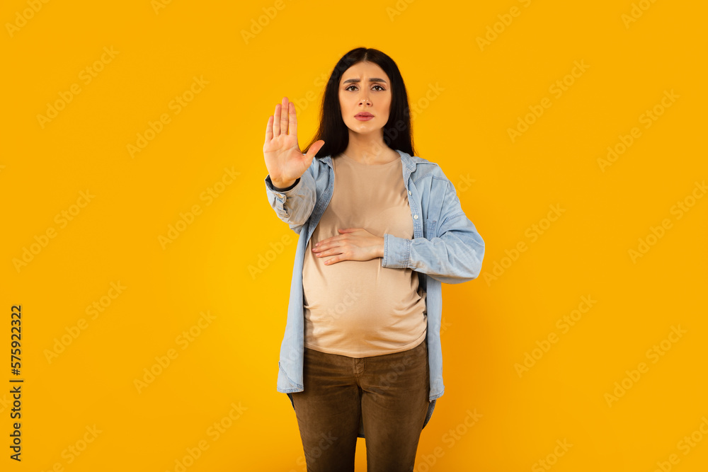 Young expectant woman doing stop sing with palm of the hand and touching belly, standing over yellow background Young expectant woman doing stop sing with palm of the hand and touching belly, standing over yellow background