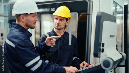 Two Caucasian production engineers in safety wear are discussing to plan for operating CNC machine in the factory. Male factory workers are controlling the process of production during work hours.