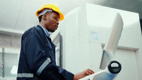 African production engineer in safety wear is operating CNC machine in the factory. A male factory worker with responsibility for programming industrial machine to control the process of production.