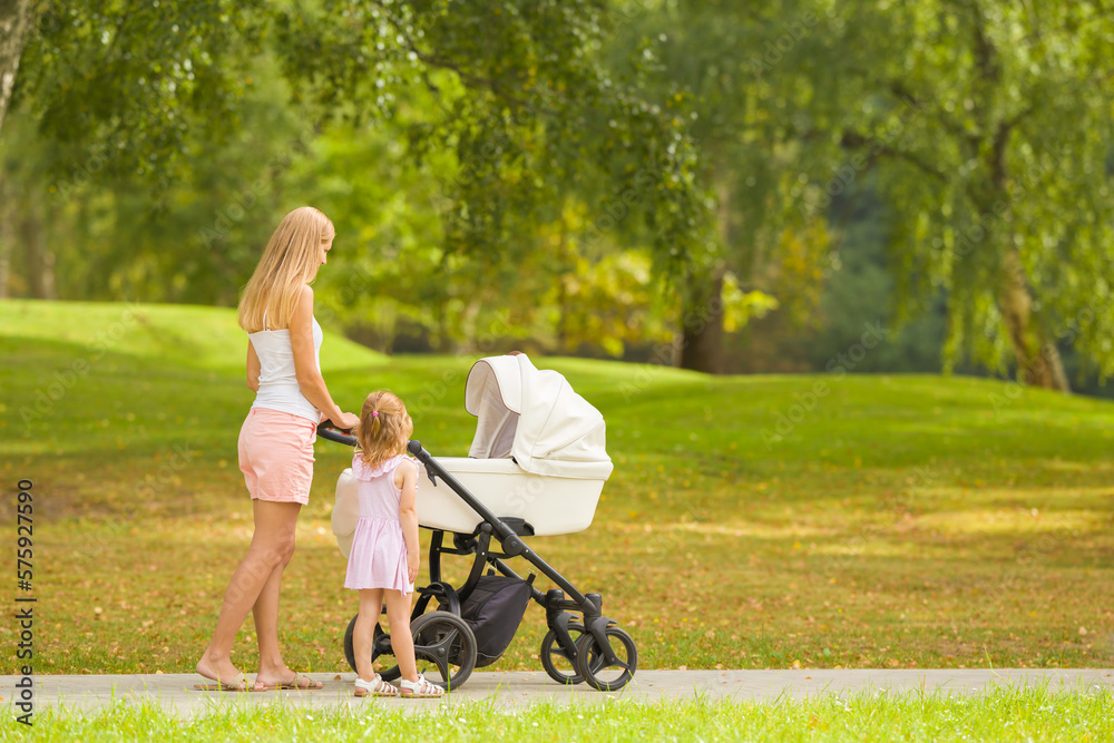Young adult mother and little daughter pushing white baby stroller and walking at town park in warm sunny summer day. Spending time together and breathing fresh air. Two child mom. Side view. Young adult mother and little daughter pushing white baby stroller and walking at town park in warm sunny summer day. Spending time together and breathing fresh air. Two child mom. Side view.