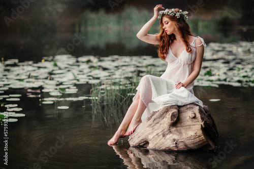 Fotografie Beautiful red haired girl in white vintage dress and wreath of flowers sitting on the shore of river