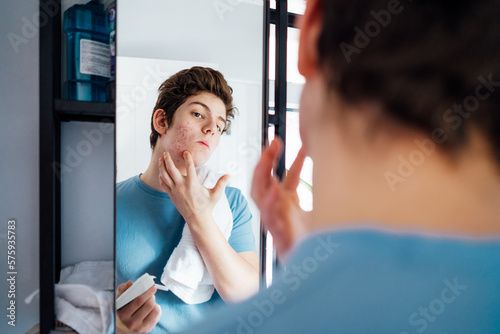 Concentrated caucasian teenage boy with acne problem take care his face skin at home. He looking in mirror and aplying cream on the face in bathroom. Teenager skin care every day treatment process photo