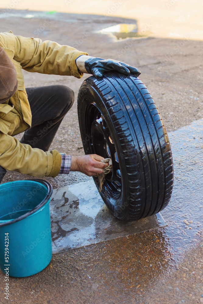 Fototapeta premium Outdoors, a man in a yellow dirty jacket with a hand without a glove washes a wheel from a car with a rag and water from a bucket, rinses the cracks, drops drip onto the mat, copy space.