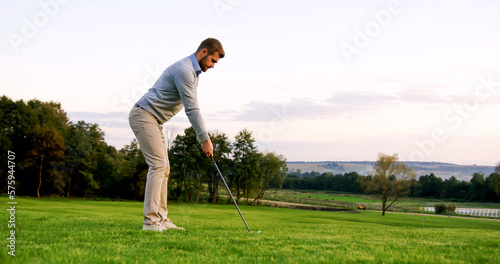 Attractive Caucasian man hitting a ball with a club on a good sunny weather day. Outdoor. Golf field.