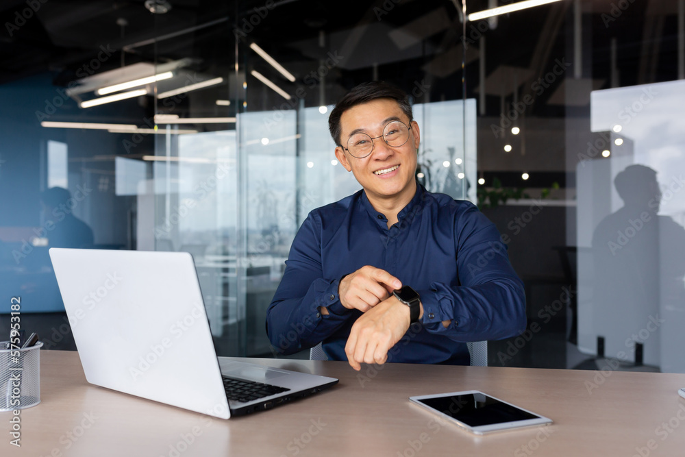 Portrait of successful smiling asian man inside office, businessman smiling and looking at camera, pointing finger at smart watch, man at workplace with laptop, programmer coding software. Portrait of successful smiling asian man inside office, businessman smiling and looking at camera, pointing finger at smart watch, man at workplace with laptop, programmer coding software.
