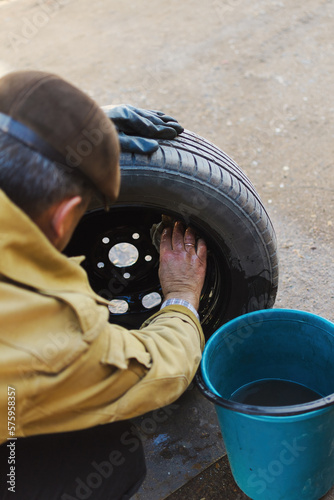 Rear view from behind the back of a man in a yellow jacket, who washes the wheel from the car with a rag and water, washing off dirt, there is a bucket next to it, copy space.