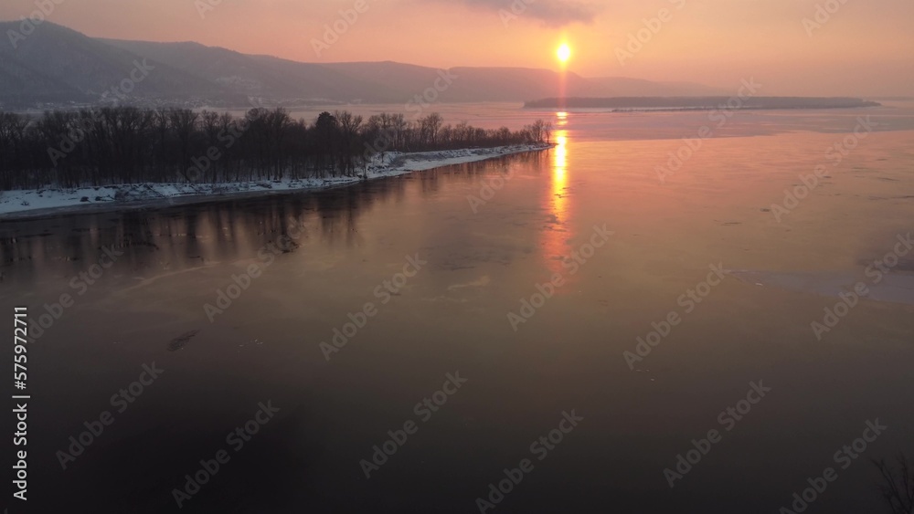View of the hills by the river at sunset. Orange sun over my river. Snow lies on the island. Top view from a drone.