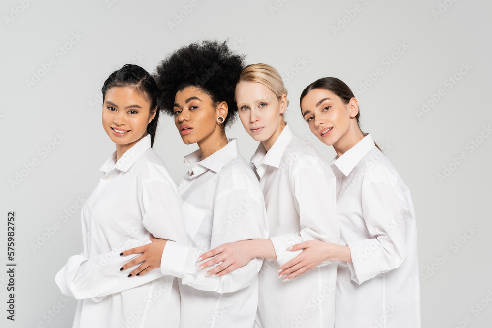 young multiethnic women looking at camera while posing in white shirts isolated on grey. young multiethnic women looking at camera while posing in white shirts isolated on grey.