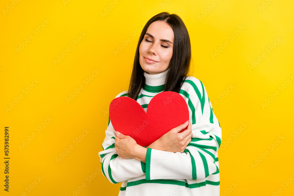 Photo of dreamy adorable woman wear striped sweater embracing red heart empty space isolated yellow color background Photo of dreamy adorable woman wear striped sweater embracing red heart empty space isolated yellow color background