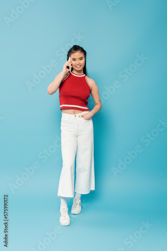 full length of positive asian woman posing with hand in pocket of white pants and talking on smartphone on blue background.