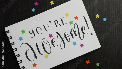 YOU'RE AWESOME! lettering in notebook with colorful stars on black wooden desk