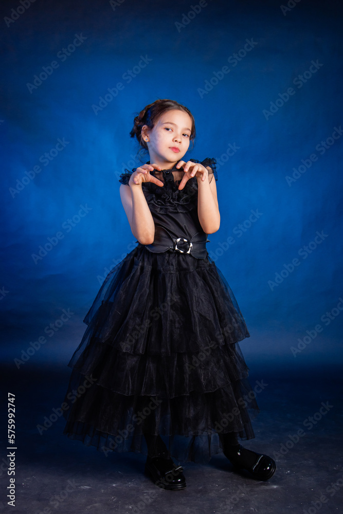 A little girl in a black dress with a pigtail hairstyle on her head poses, isolated on a dark background with blue backlight.