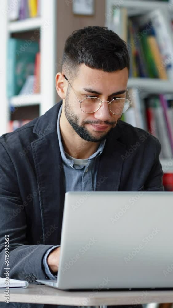 Vertical Screen: Handsome young man with eyeglasses sitting in library and browsing internet on laptop, looking stressed and unhappy. Distance learning. Remote work. Concept of technology Vertical Screen: Handsome young man with eyeglasses sitting in library and browsing internet on laptop, looking stressed and unhappy. Distance learning. Remote work. Concept of technology
