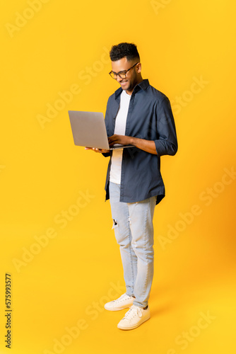Full length photo of charismatic indian man is standing and typing on the laptop, handsome guy in casual wear working remotely, responding emails, copy space