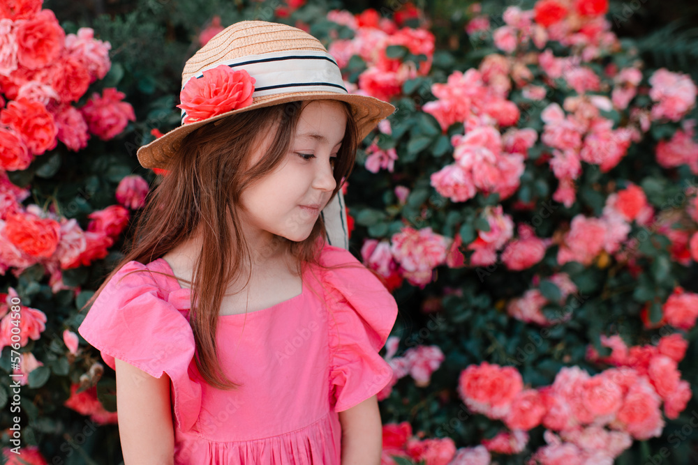 Smiling cute child girl 7-8 year old wear straw hat and pink dress posing over blooming rose flowers in garden outdoor. Summer season. Childhood.