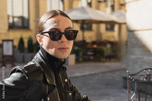 tattooed young man in stylish sunglasses and black leather jacket looking at camera outdoors.