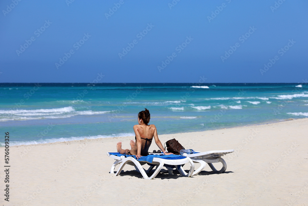 Girl in swimsuit tanning on deck chair on sea waves background. Beach with white sand on Atlantic ocean coast