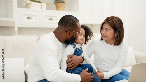 Multiracial newbie parent is sitting on a sofa with their daughter and giving love to her while watch online entertainment contents on a laptop. African adult man is kissing his daughter to show love. © Benixs