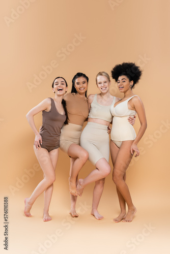 full length of young and happy multiethnic women in lingerie embracing and looking at camera on beige background.