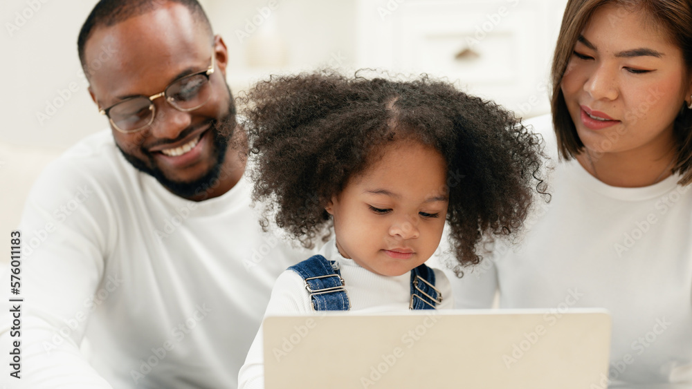 A cheerful daughter focus on looking a laptop screen to watch online kid content with multiracial newbie parent take care of her. A happy family is spending time to watch online entertainment content. A cheerful daughter focus on looking a laptop screen to watch online kid content with multiracial newbie parent take care of her. A happy family is spending time to watch online entertainment content.