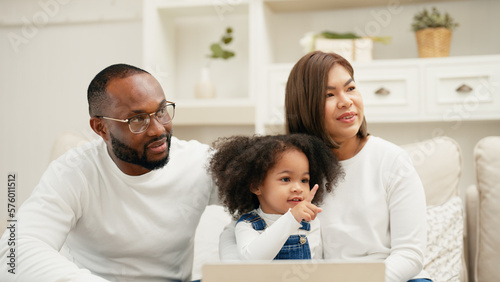 Multiracial newbie parent is sitting on a sofa with their daughter and watching online entertainment contents on their laptop. A cheerful daughter is talking to her father and mother with curiosity.