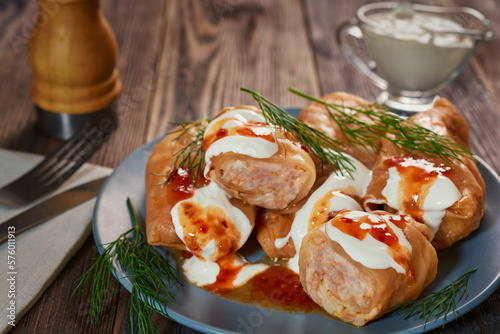 Cabbage rolls poured with sour cream and sauce lie in a small gray plate, which stands on a dark wooden table.