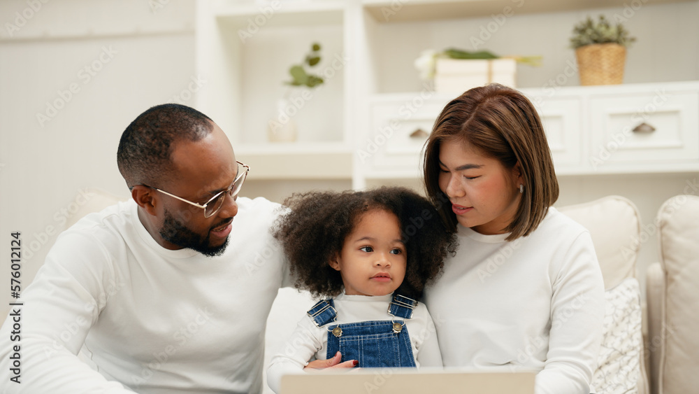 Multiracial newbie parent is sitting on a sofa with their daughter and watching online entertainment contents on their laptop. A cheerful daughter is talking to her father and mother with curiosity. Multiracial newbie parent is sitting on a sofa with their daughter and watching online entertainment contents on their laptop. A cheerful daughter is talking to her father and mother with curiosity.