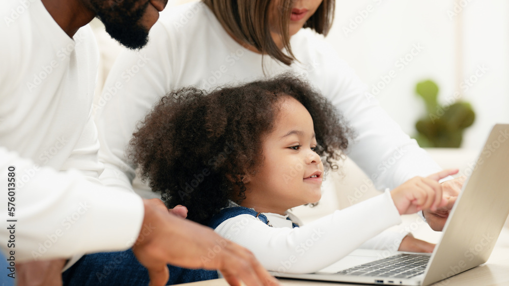 Obraz premium Close-up of a curious daughter touching a laptop screen to select online kid contents while multiracial newbie parent taking care of her. A happy family is teaching their daughter to use a laptop.