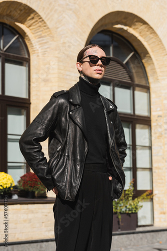 tattooed man in stylish sunglasses and black leather jacket standing with hands in pockets on urban street.