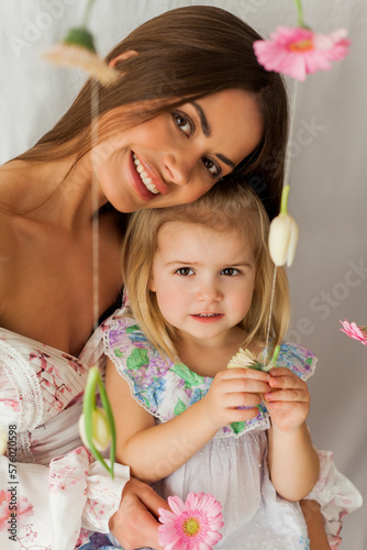 Beautiful young long-haired mother with little daughter of 3 years old with colored flowers on white background. Mothers Day. Holidays. Happy child.