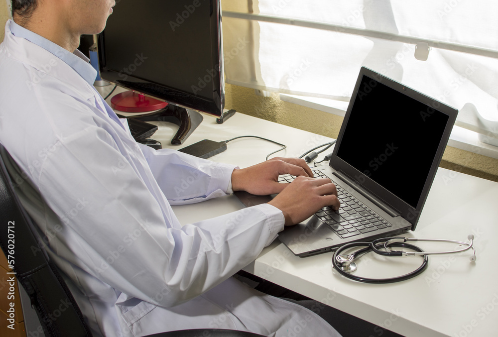 A doctor is sitting at a desk with a laptop and a monitor 