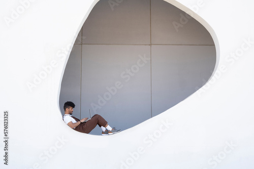 Man using laptop sitting on wall with hole in building
