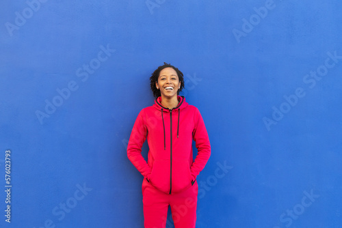 Happy woman with hands in pockets leaning on blue wall