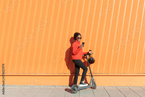 Woman using smart phone with electric push scooter in front of orange wall photo