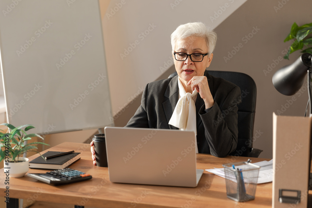 Confident stylish european middle aged senior woman using laptop at workplace. Stylish older mature 60s gray haired lady businesswoman sitting at office table. Boss leader teacher professional worker Confident stylish european middle aged senior woman using laptop at workplace. Stylish older mature 60s gray haired lady businesswoman sitting at office table. Boss leader teacher professional worker