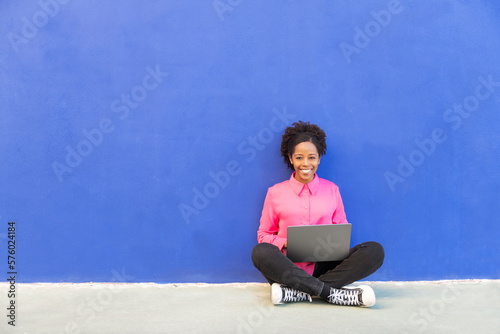 Happy woman with laptop sitting cross-legged in front of wall