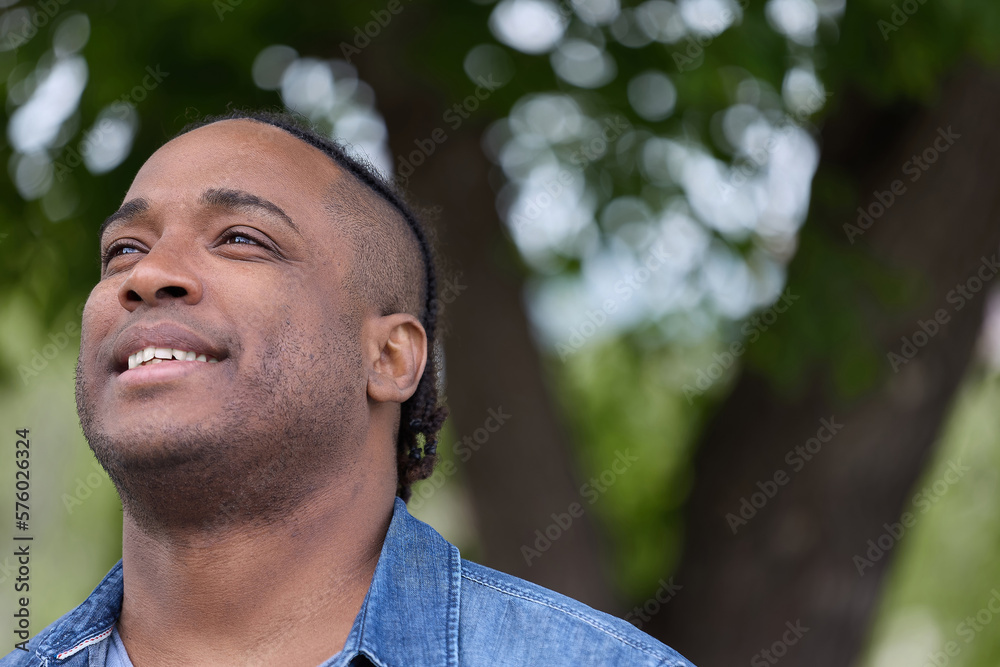 Happy African-born attractive man in jeans dreams of standing under a tree in a park. Romantic African-American in Spring City Park Looks Into the Distance Happy African-born attractive man in jeans dreams of standing under a tree in a park. Romantic African-American in Spring City Park Looks Into the Distance