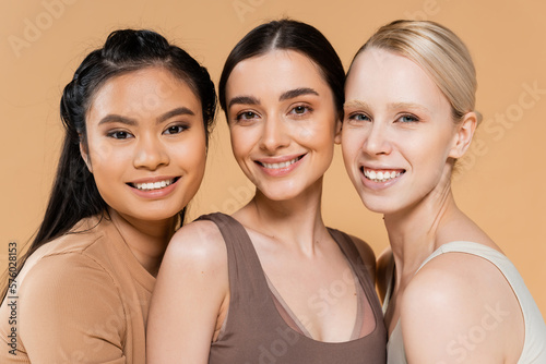 pretty multiethnic women in lingerie smiling at camera isolated on beige.